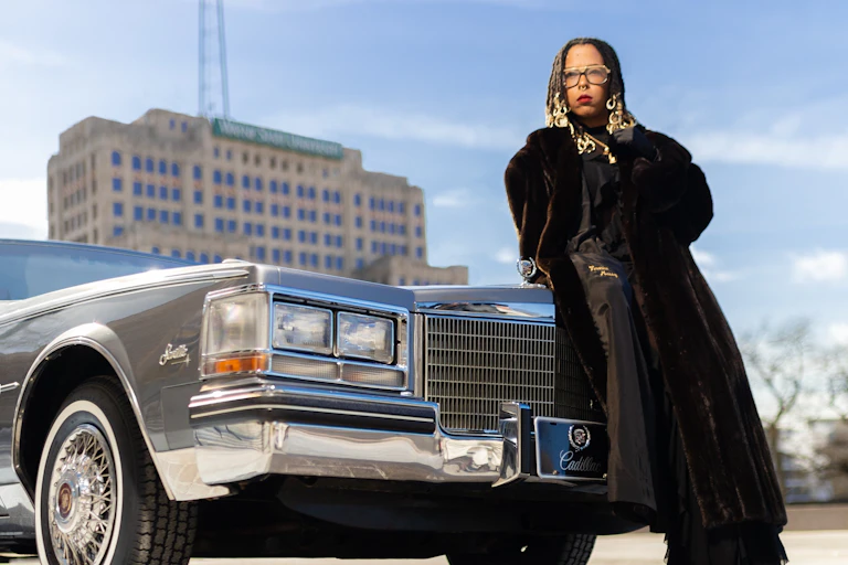 A Black woman wearing a long black coat, glasses and red lipstick leans against a Cadillac