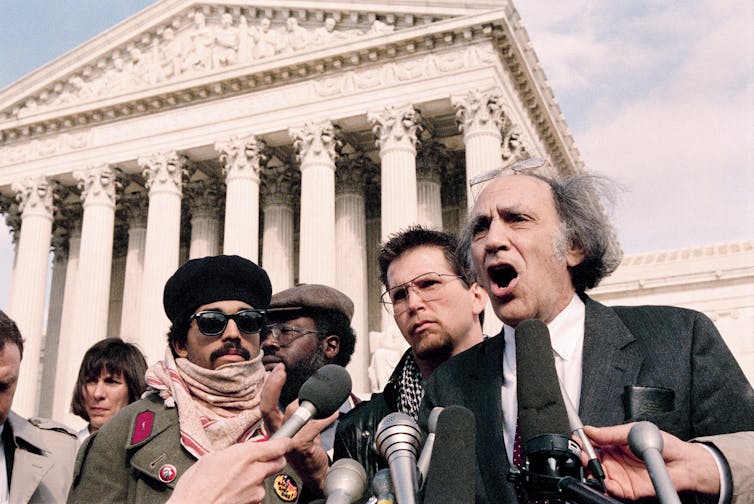 A man speaks at a podium in front of the Supreme Court building, surrounded by other men