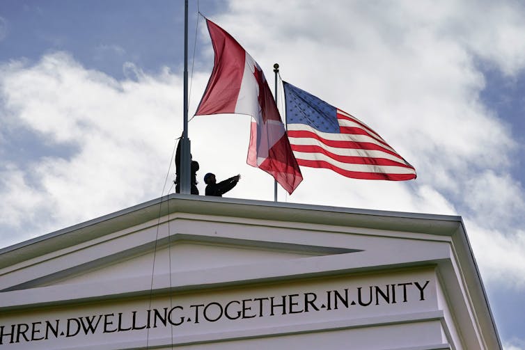 Workers raise a Canadian flag next to an American one on top of a stone arch that reads Dwelling Together in Unity.