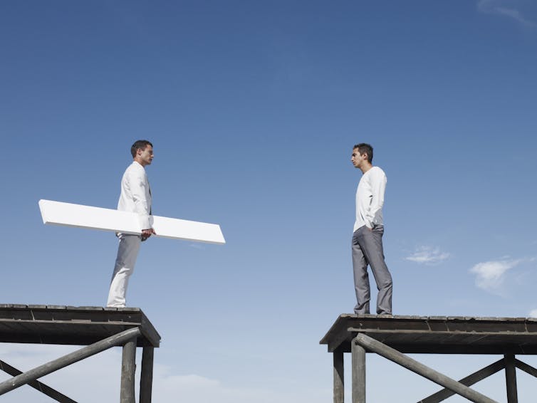 Two men on elevated stages separated by a gap. One man holds a plank that could span the distance.