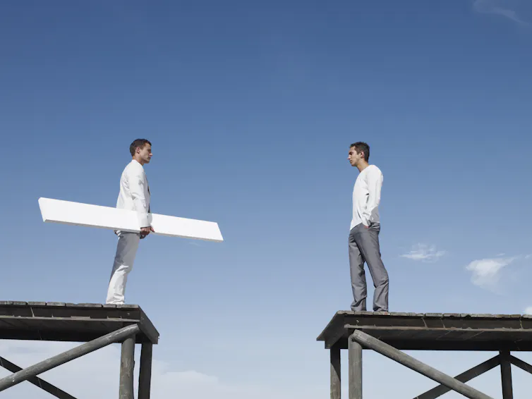 Two men on elevated stages separated by a gap. One man holds a plank that could span the distance.