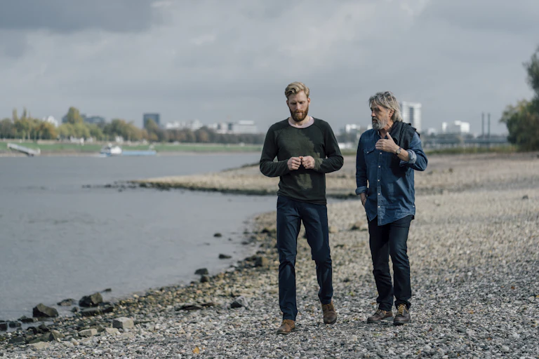 Two men walk on a beach with a cityscape behind them.
