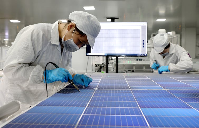 Two workers in white smocks and hats use instruments for detailed work on line of solar cells in a factory.