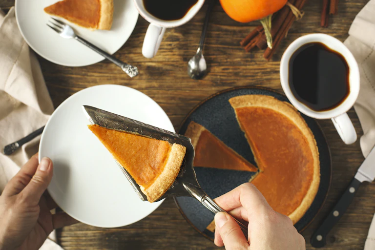 Pumpkin pie next to cups of coffee, with a person's hands shown putting a slice of pie on a plate.