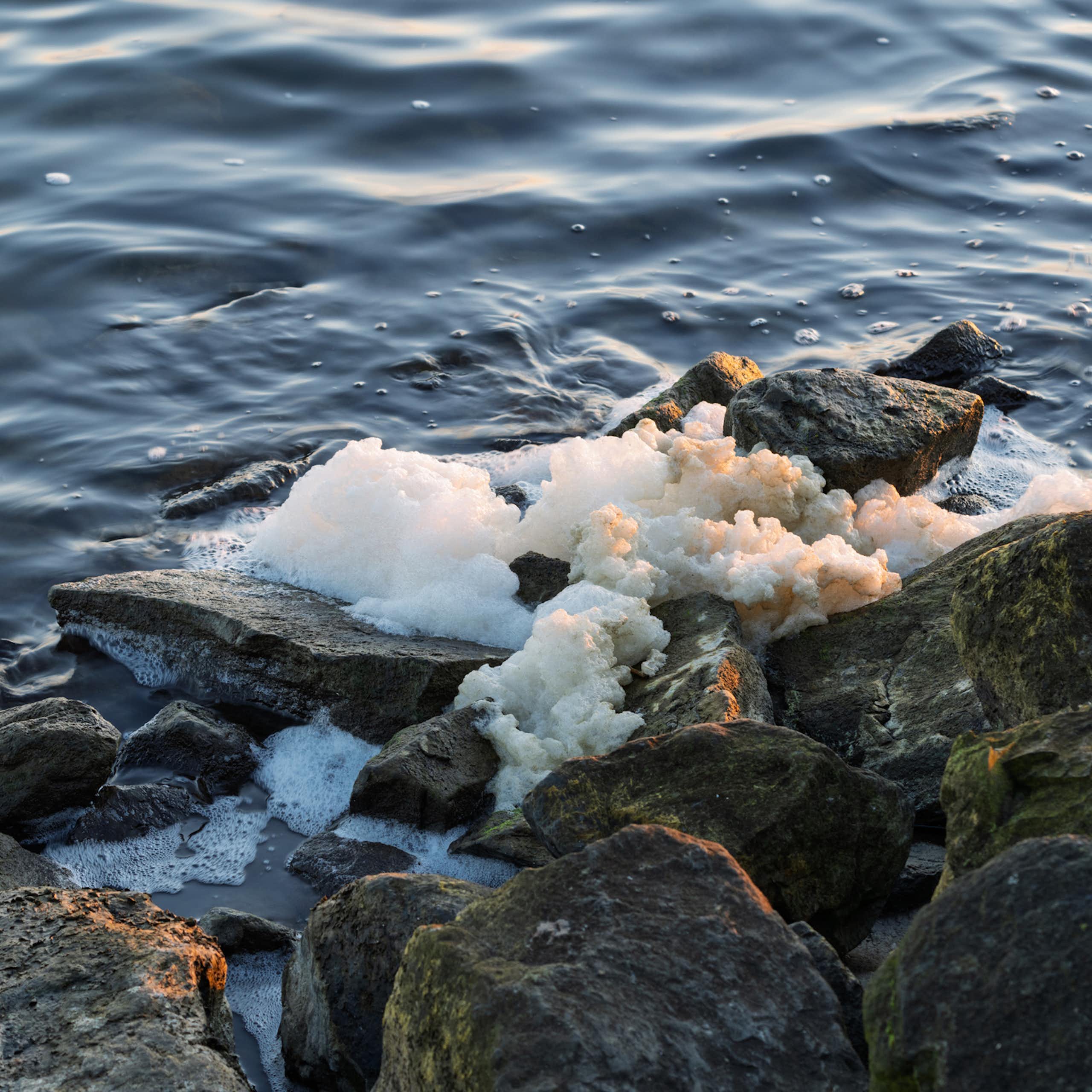 Sea foam on top of rocks by a body of water.