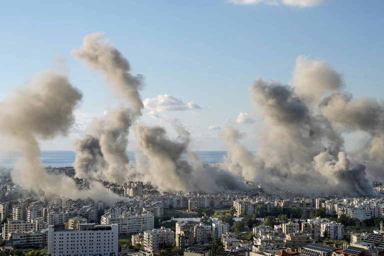 dark smoke billows from dense urban area with blue skies above and the sea in the distance