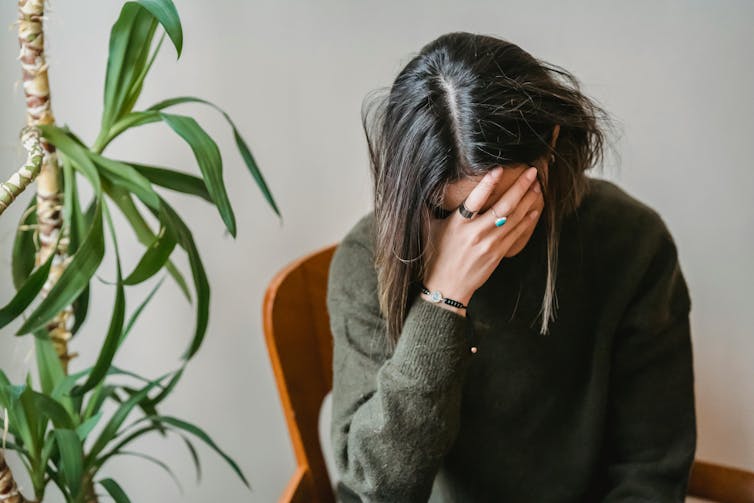A woman sits next to an indoor plant. Her head is in her hand.