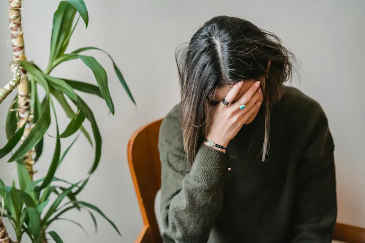 A woman sits next to an indoor plant. Her head is in her hand.
