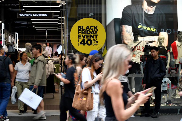 Retail shoppers walk on the street in front of a window with advertisements for a Black Friday sale