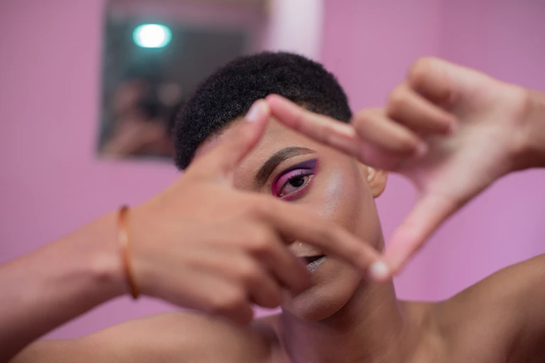 A young trans person looks through a rectangular window made with their hands.