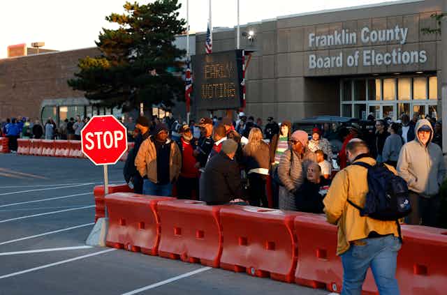 a group of people stand outside between a row of traffic barriers and the entrance to a concrete building