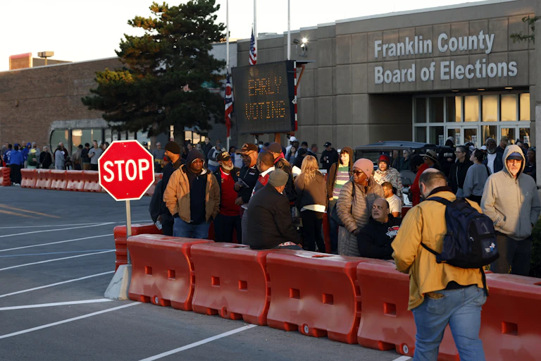 a group of people stand outside between a row of traffic barriers and the entrance to a concrete building
