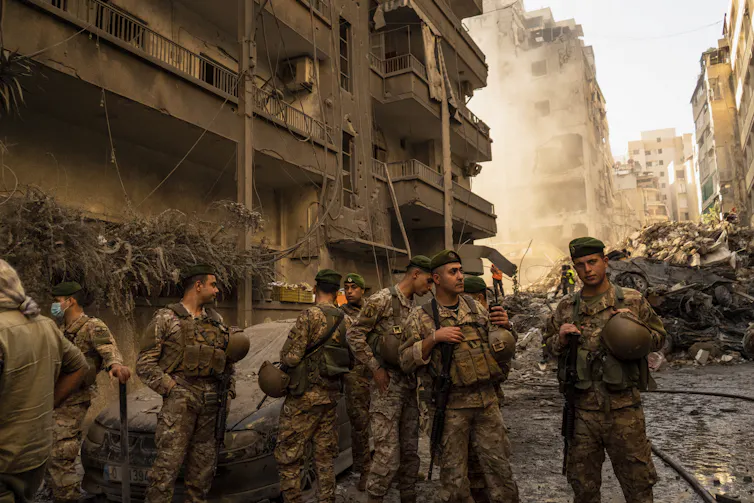 Men in army fatigues stand in the street in front of a destroyed building.