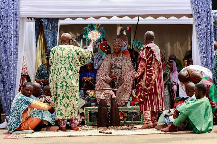An elaborate set-up with a man in traditional clothing seated on a throne in a tent with attendants and elaborately beaded sculptures.