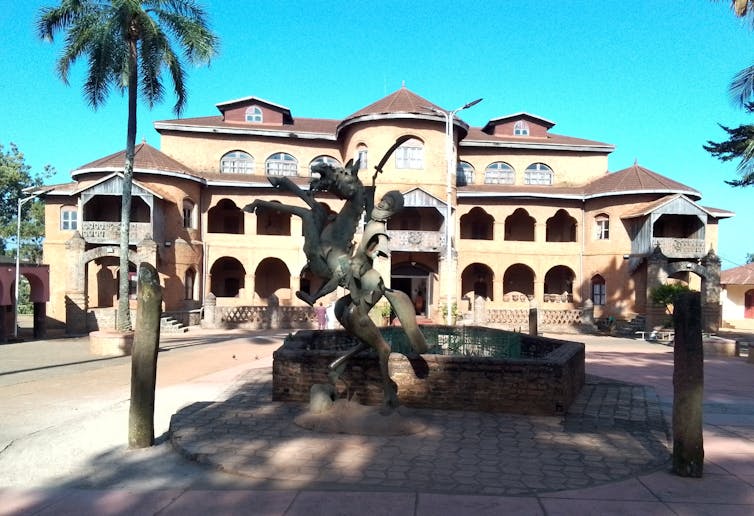 A grand old structure of multiple storeys with arches and a fountain and statue in front of it.
