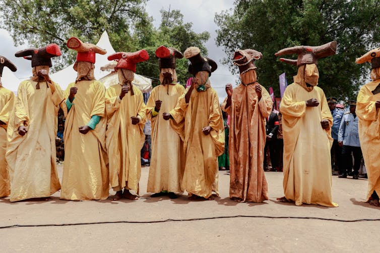 A row of people in gold gowns, their faces covered in fabric masks, on their heads replicas of animal heads.