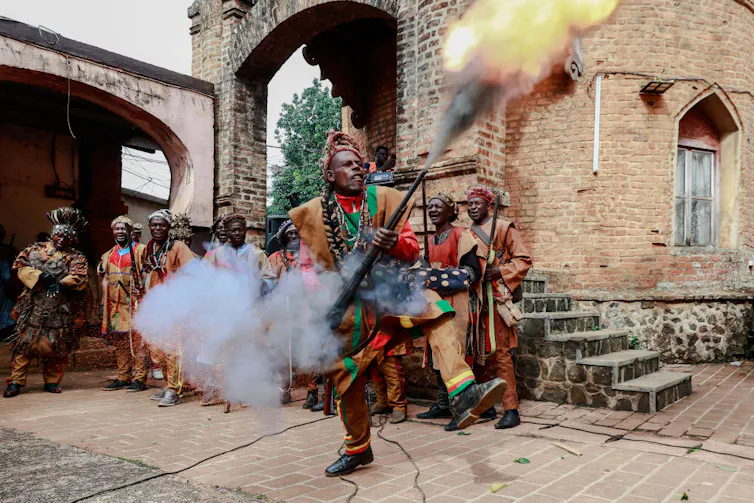 A dramatic image of a man with a foot raised fires a weapon in a traditional military outfit, others in similar attire looking on.