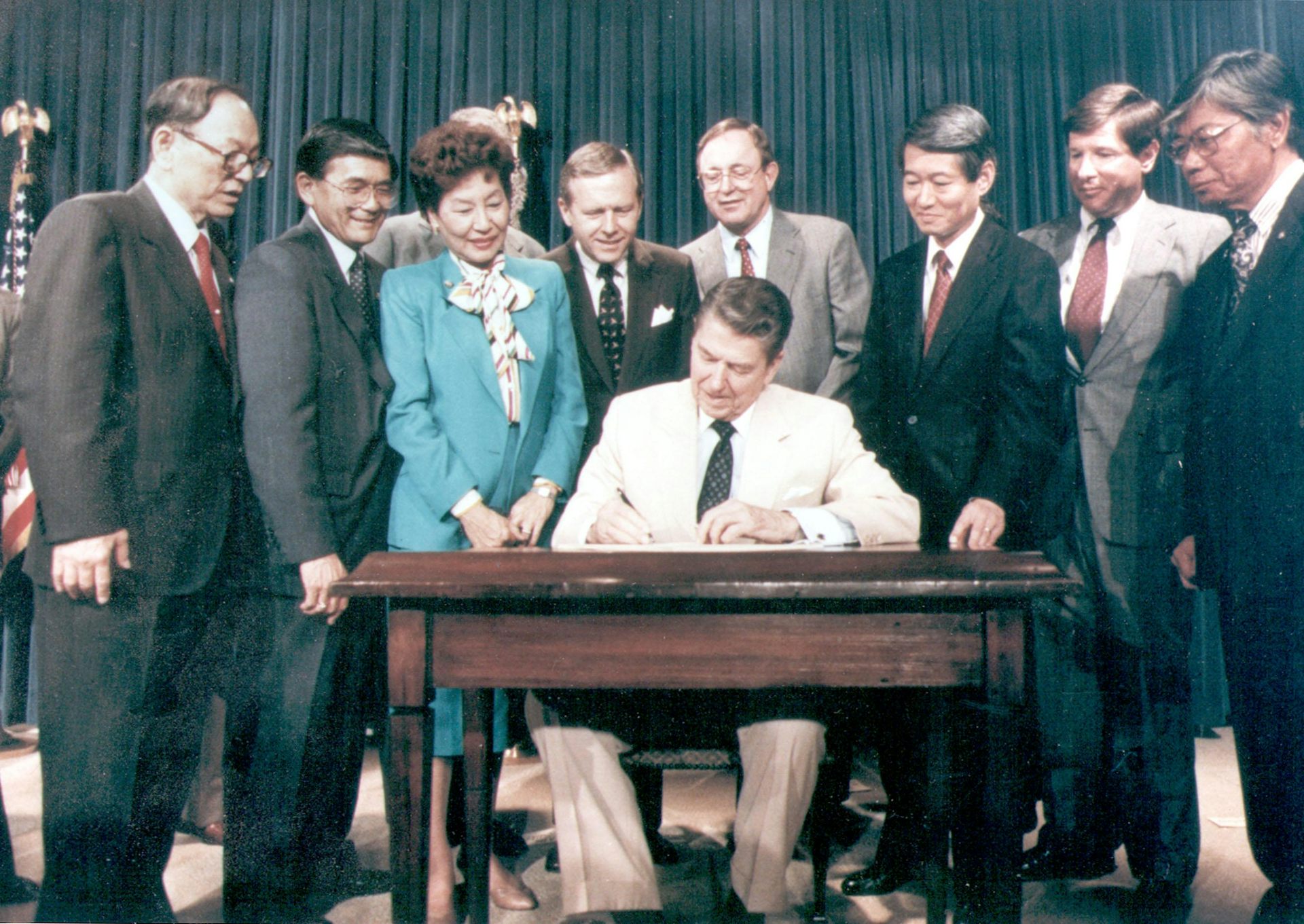 A man in a suit sits at a table surrounded by a group of other people.