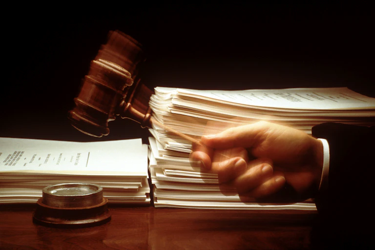 A hand swings a gavel down in front of large stacks of paper.