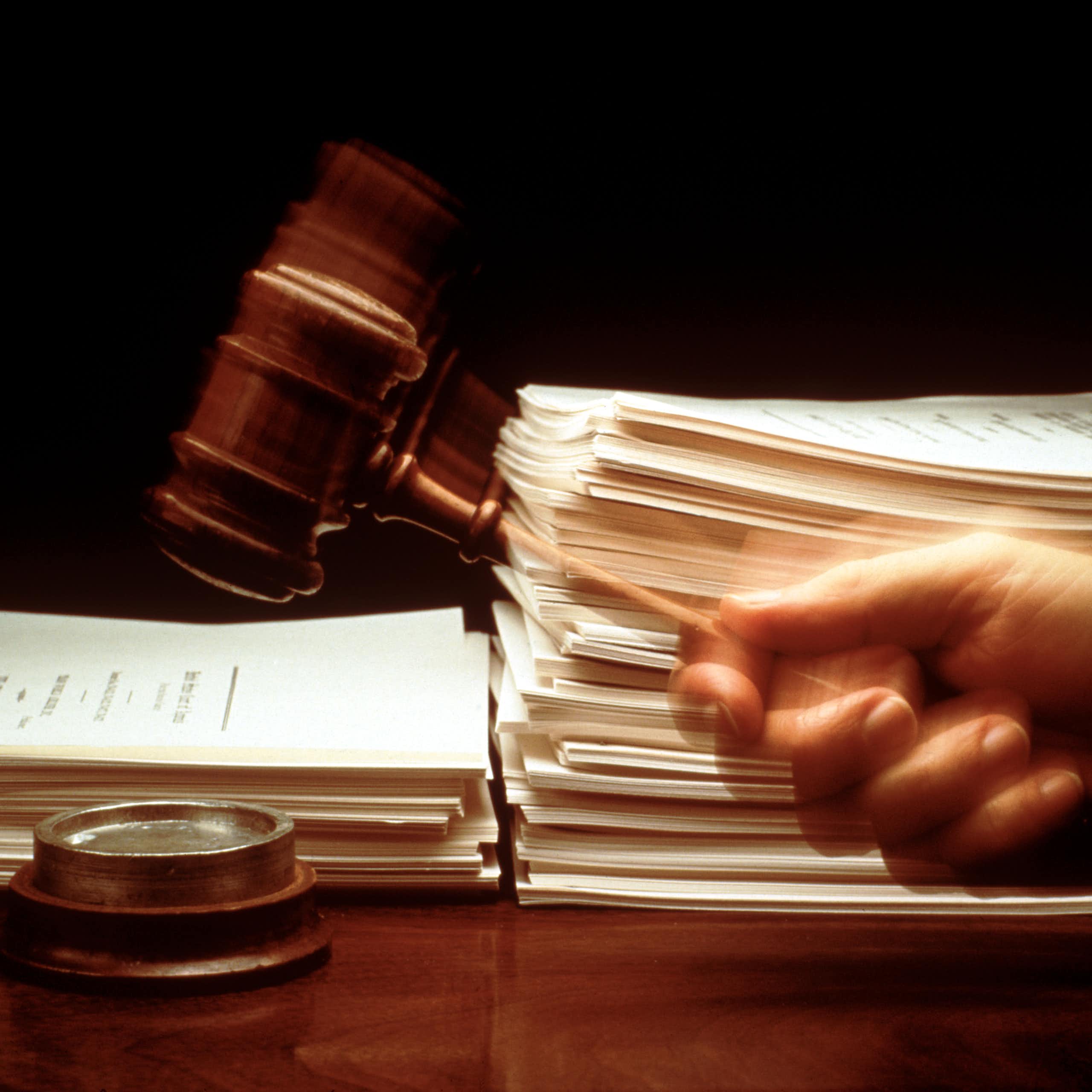A hand swings a gavel down in front of large stacks of paper.
