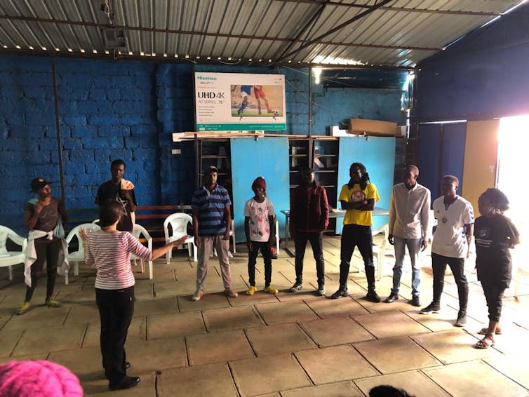 10 people standing in semi circle in darkly lit hall, talking to one person