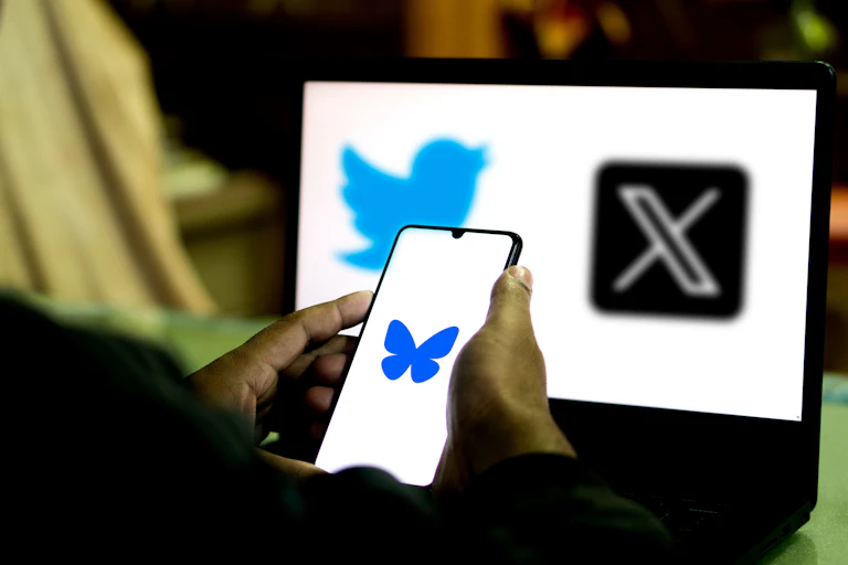 Photo of a person's hands holding a phone showing a blue butterfly logo, with a screen in the background showing the Twitter and X logos.