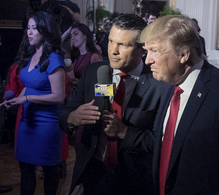 A rotund blond man with greyish blond fluffy hairs stands next to a younger dark-haired man holding a microphone.