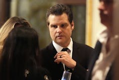 A dark-haired man in a suit and tie speaks to people around him.