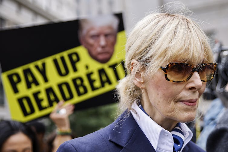 A blonde woman wearing sunglasses stands in front of a sign featuring a photo of Donald Trump that reads Pay Up Deadbeat.