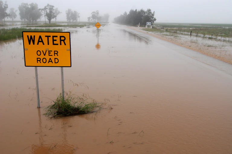 Flood waters covering a rural road in Australia, with sign "Water Over Road" visible.