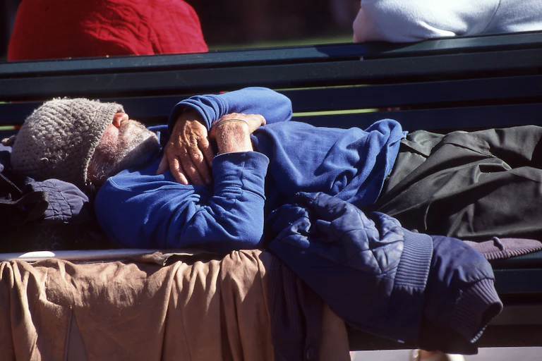 An older man lies on a bench with a beanie pulled across his eyes.