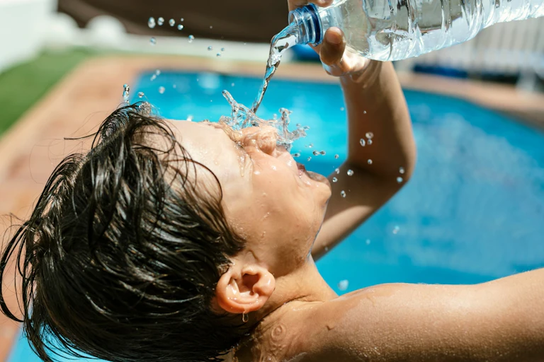 Teenage boy pouring bottled water over face, next to outside swimming pool