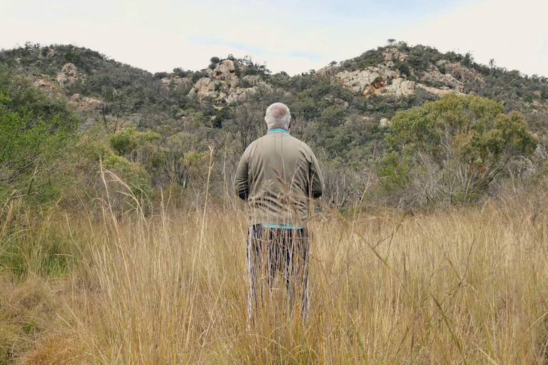 Photo of a man standing in long grass facing away from the camera, with hills in the background.