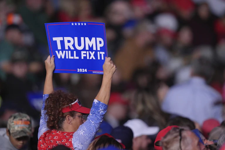 A woman in a red visor holds up a sign that reads Trump Will Fix It.