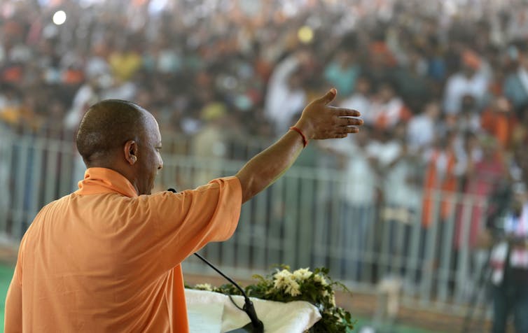 A man in orange is seen from behind raising his right hand to a crowd.
