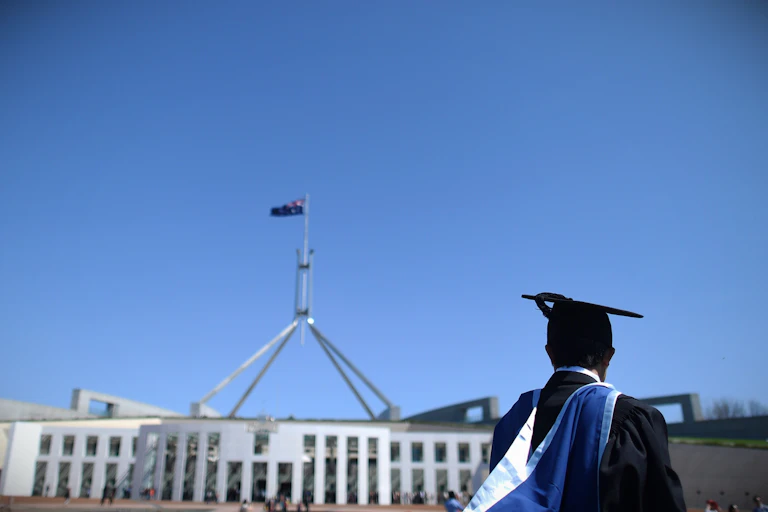 A university graduate in cap and robe stands in the forecourt of Parliament House in Canberra.