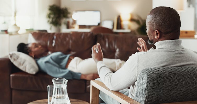 A male therapist talks to a woman lying on a couch with her eyes closed.
