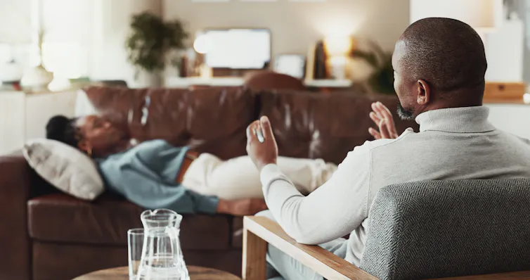 A male therapist talks to a woman lying on a couch with her eyes closed.