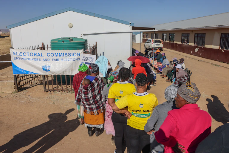 Voters, mainly women, wait to cast their votes. Some are standing, others are sitting on chairs.