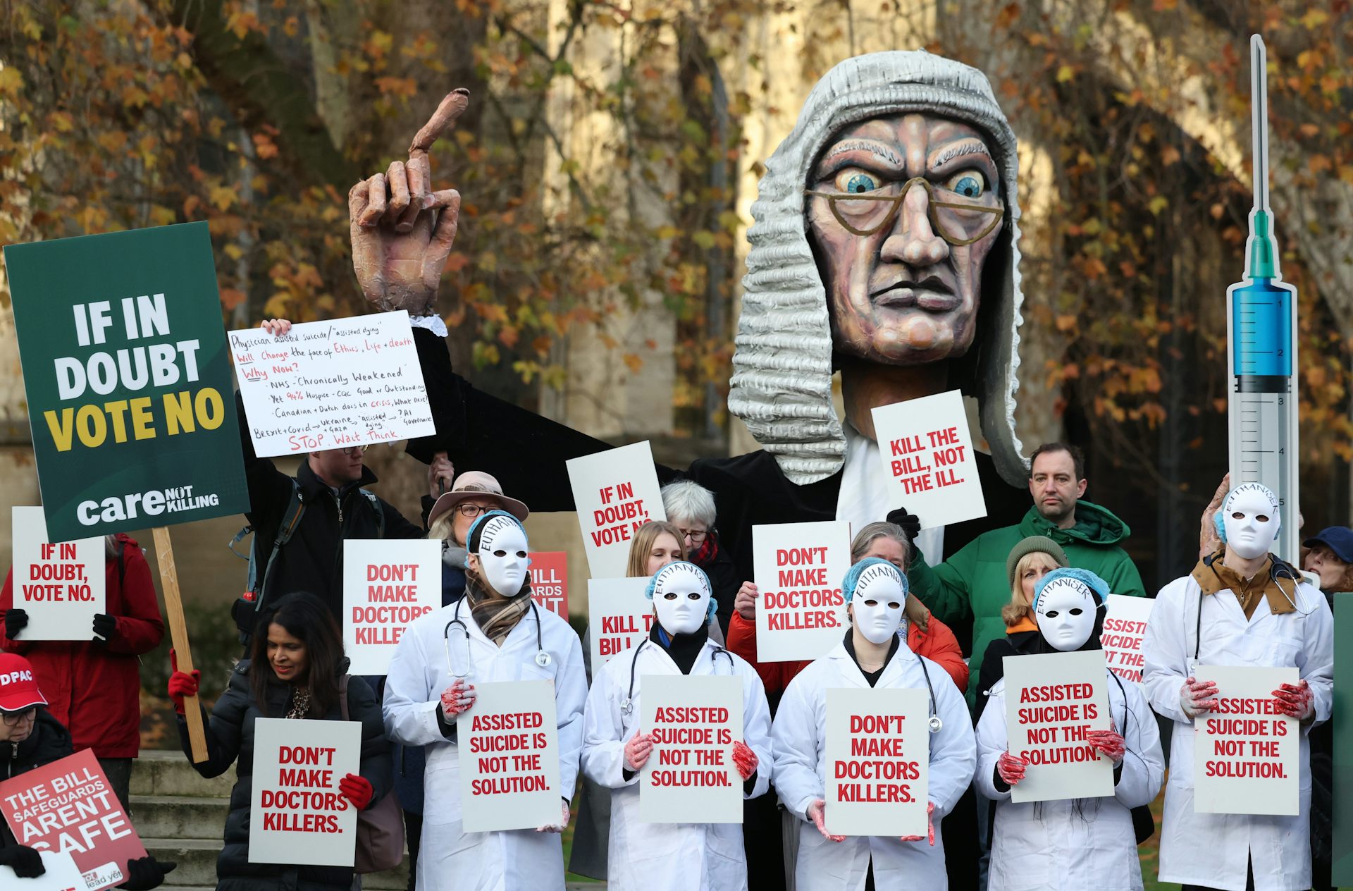 Os manifestantes com jalecos médicos brancos e máscaras brancas, com tinta vermelha nas mãos, seguram cartazes com slogans como “não faça dos médicos assassinos”. Uma grande efígie de um juiz furioso apontando o dedo aparece atrás deles.
