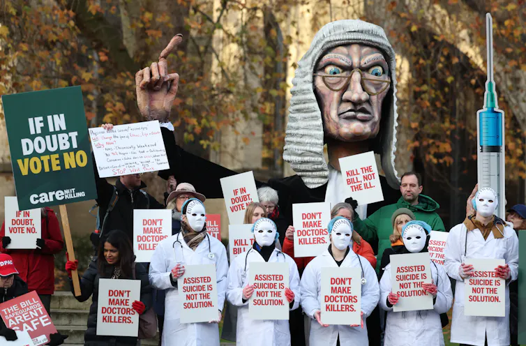 Protesters in white doctor coats and white masks, with red paint on their hands, hold signs with slogans like 'don't make doctors killers'. A large effigy of an angry looking judge pointing his finger looms behind them.