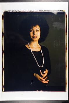 A serene black woman looking directly at the camera wearing a black dress and a string of beads.