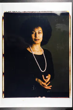 A serene black woman looking directly at the camera wearing a black dress and a string of beads.