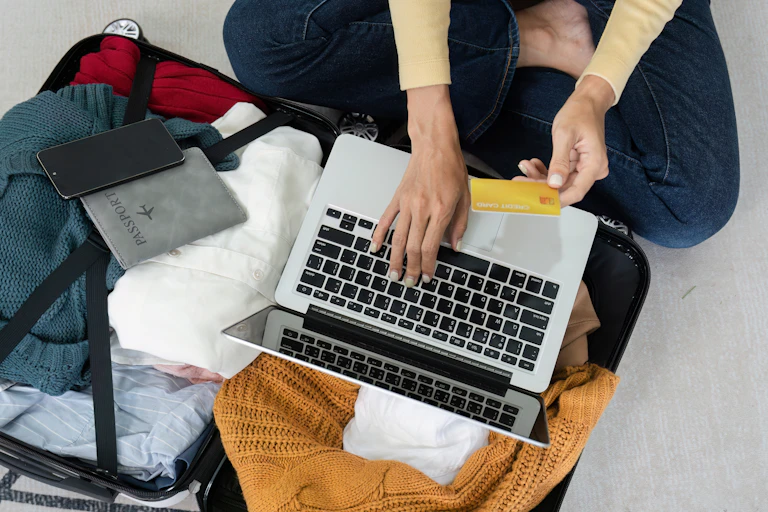 Young woman booking accommodation online and paying with credit card while sitting on floor with open suitcase