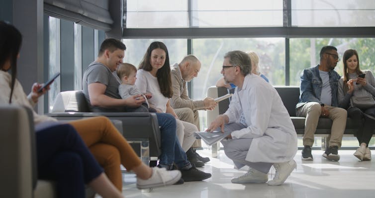 A doctor squats down talking to a toddler on a man's lap in a busy waiting room.