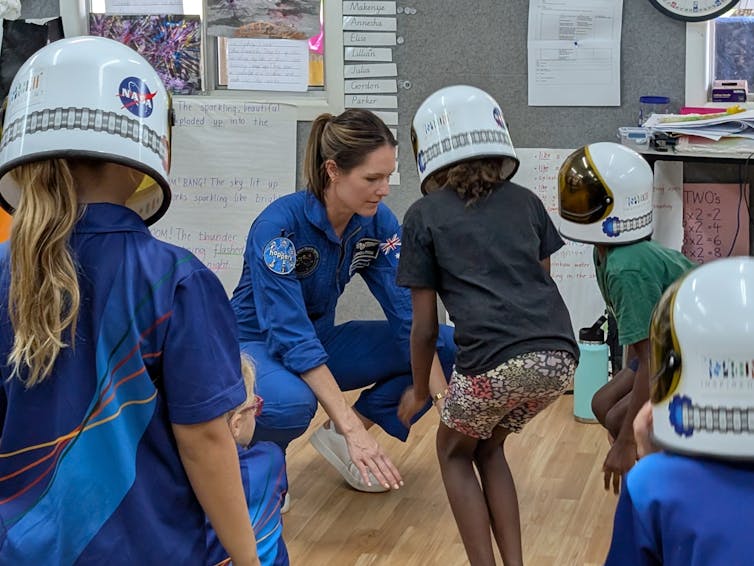 Woman wearing astronaut uniform conducts an exercise with a group of children in a classroom.