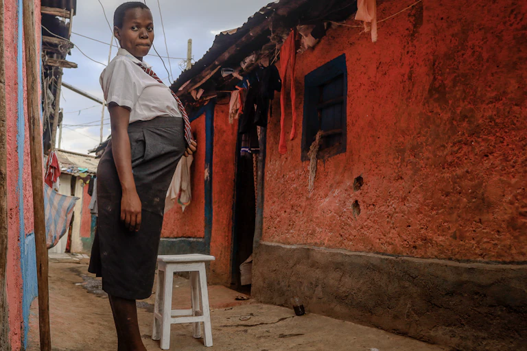 Young woman in white shirt, striped tie and grey skirt, with pregnant belly, standing outside a mud house