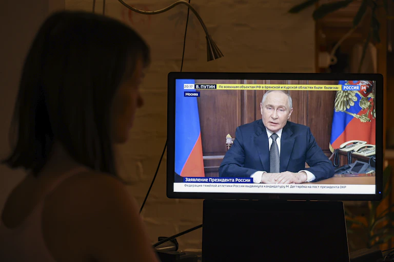 A woman watches Vladimir Putin speak on a TV screen.
