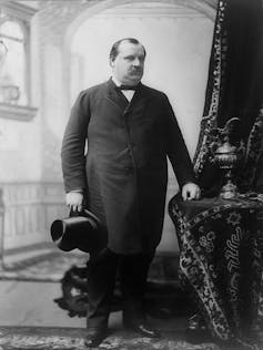 A rotund man with a moustache and in a tight coat holds his top hat in his right hand as he poses for a photo.