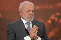 An older man with grey thinning hair and a beard clasps his hands in front of him.